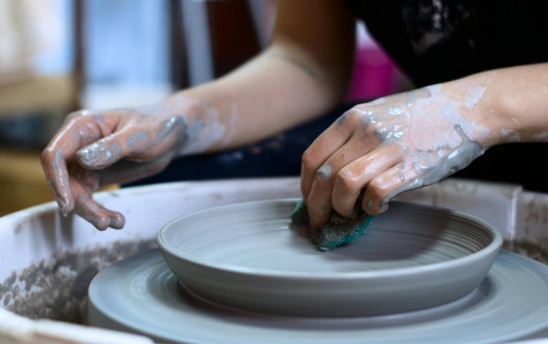 Clay-covered hands smoothing a gray stoneware bowl with a sponge on a pottery wheel